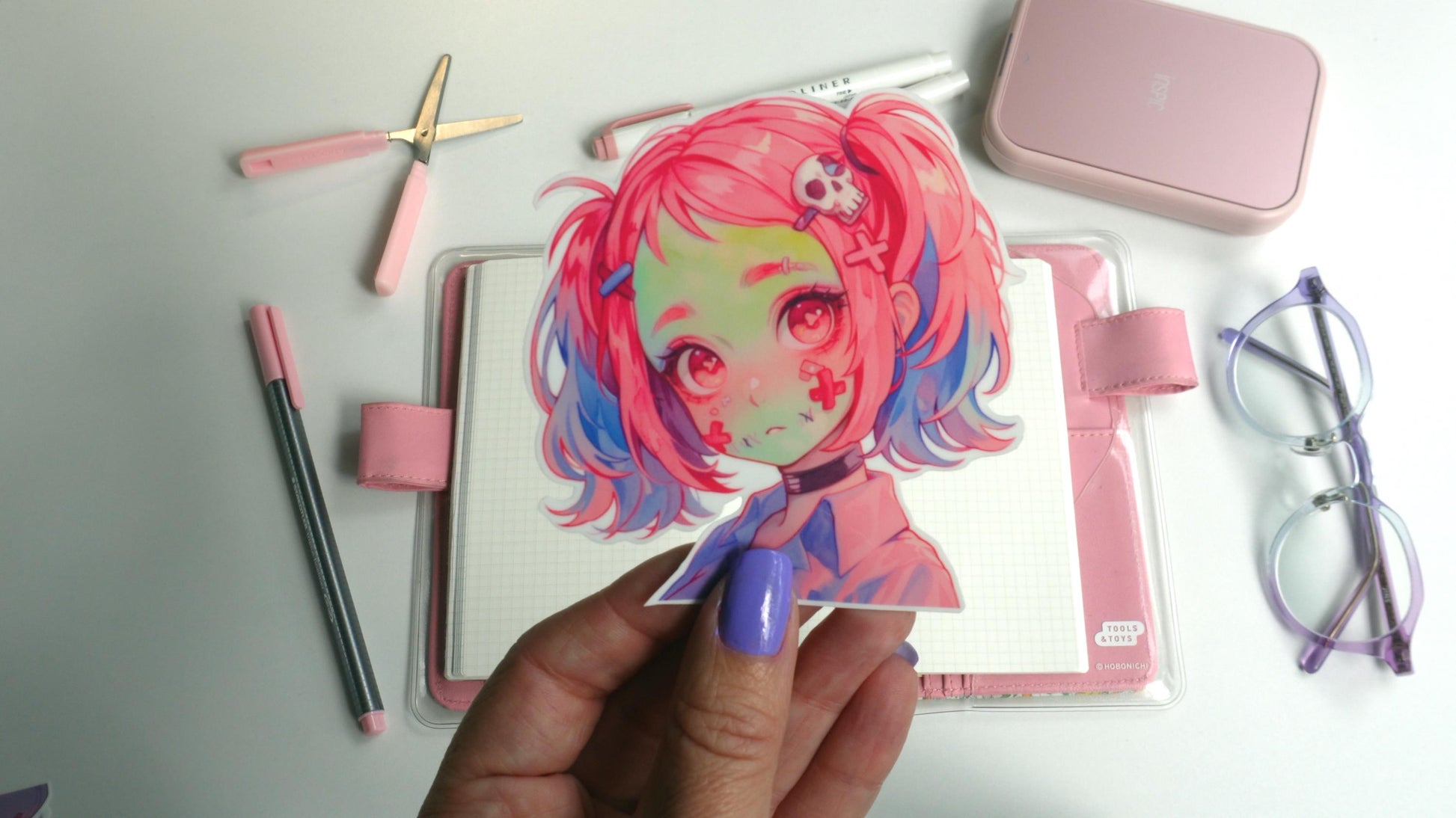 A hand with purple nails holds a sticker of an anime character with pink and blue hair, red eyes, and skull accessories. Background items include a pink notebook, pens, scissors, a case, and glasses on a white surface.