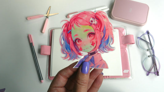 A hand with purple nails holds a sticker of an anime character with pink and blue hair, red eyes, and skull accessories. Background items include a pink notebook, pens, scissors, a case, and glasses on a white surface.