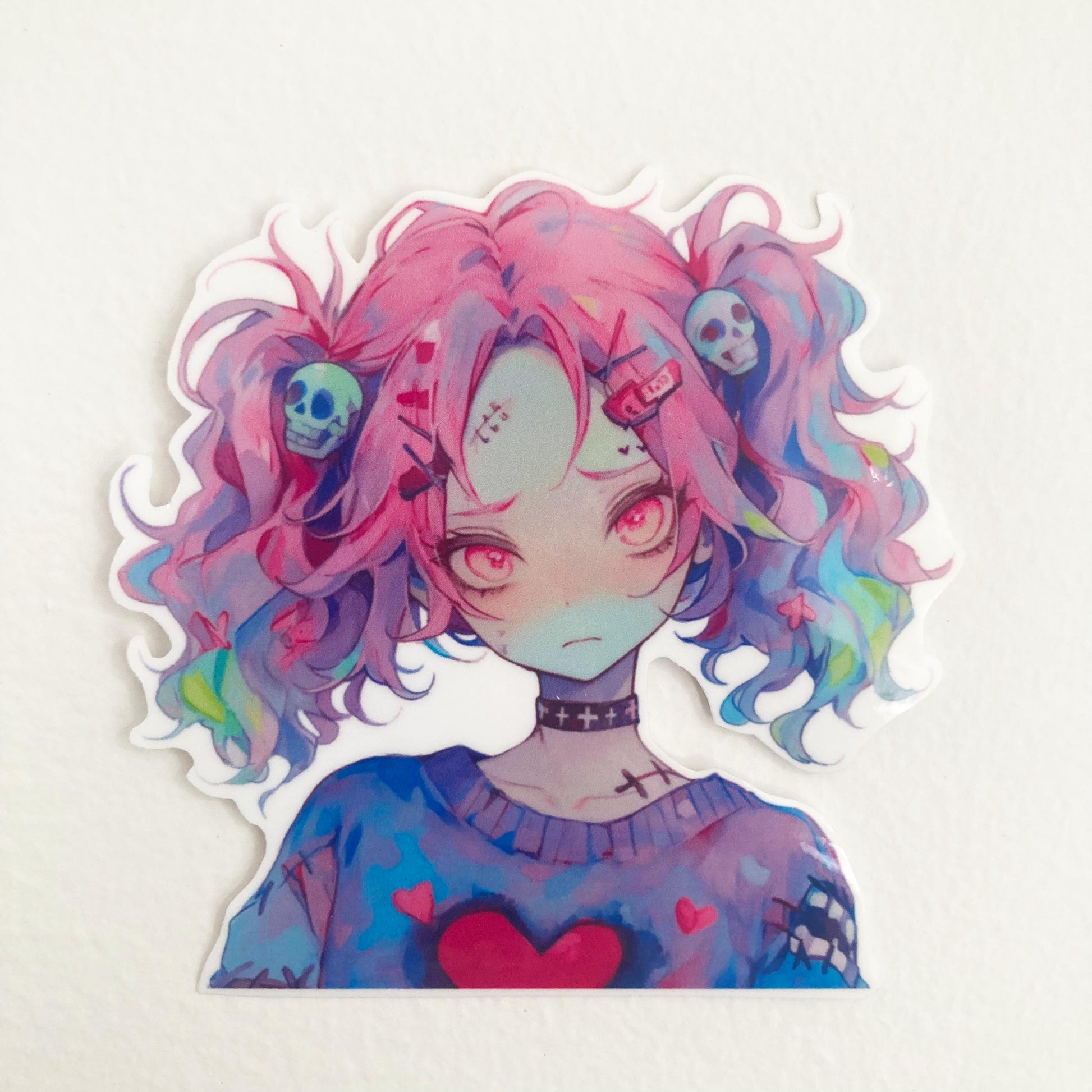 A sticker of an anime character with pink and multicolored hair, skull accessories, and a heart on their sweater, placed on a plain, light background.