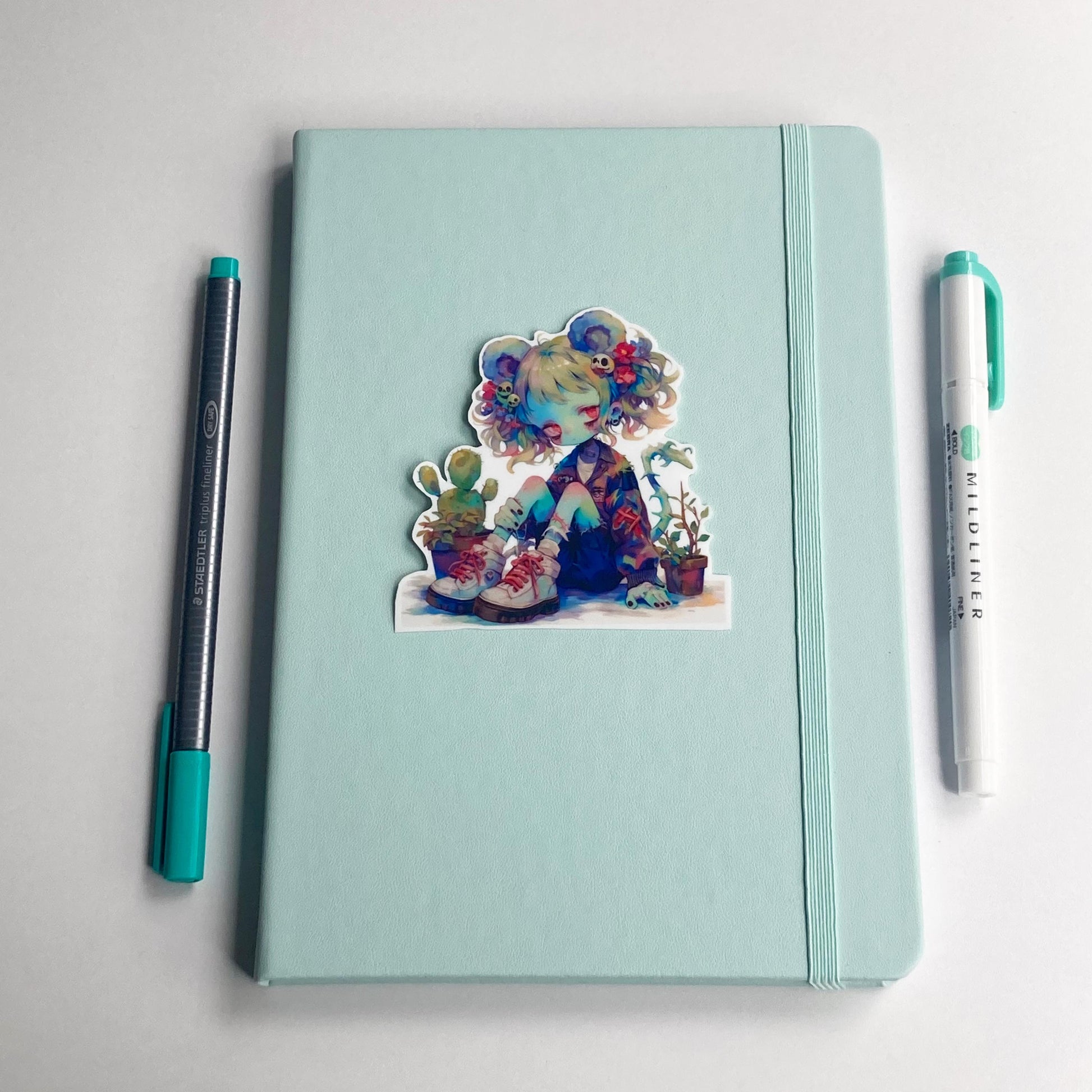 A sticker of an anime character with colorful hair, mouse ears, and potted plants is placed on a closed light teal notebook. A pen with a teal cap is on the left, and a teal marker is on the right.