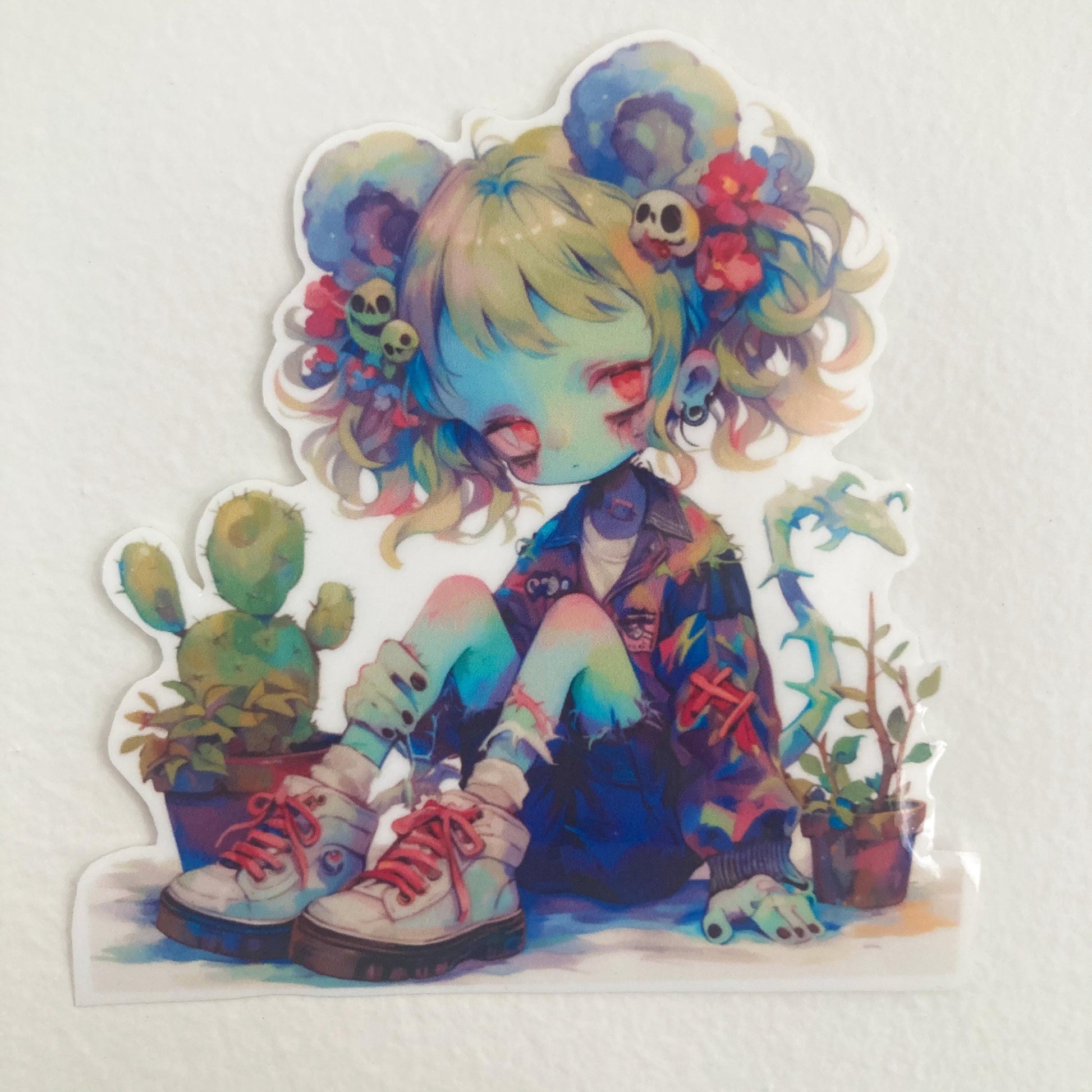 A sticker of an anime character with colorful hair, mouse ears, skull accessories, and potted plants. The character is sitting with eyes closed, wearing a jacket and sneakers. The background is a plain light surface.
