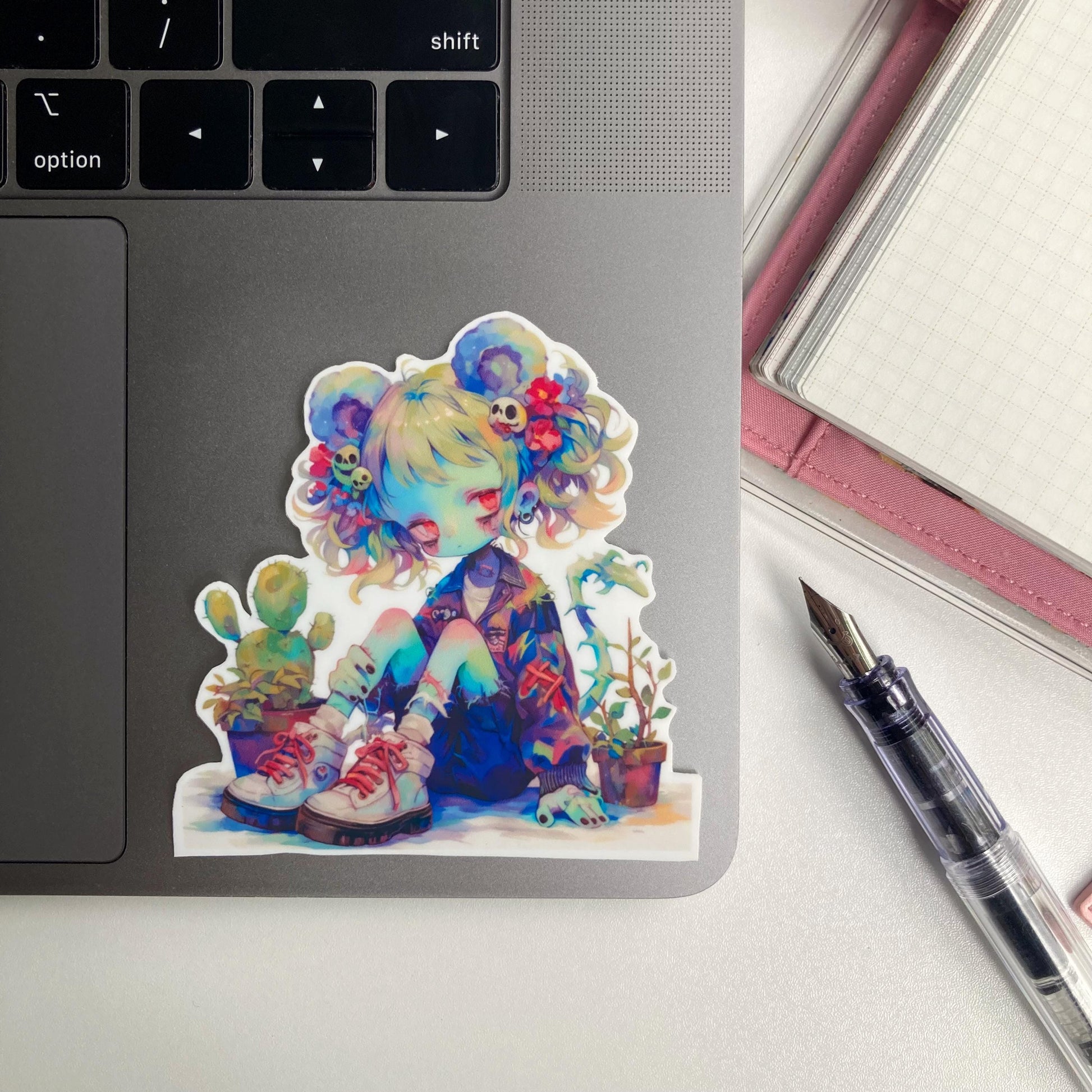 A sticker of an anime character with colorful hair, mouse ears, and potted plants is placed on a laptop. A pink notebook and a fountain pen are nearby.