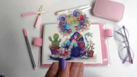 A hand with purple nails holds a sticker of an anime character with colorful hair, mouse ears, and plants. The character wears a jacket and sneakers. Background items include a pink notebook, pens, scissors, a case, and glasses on a white surface