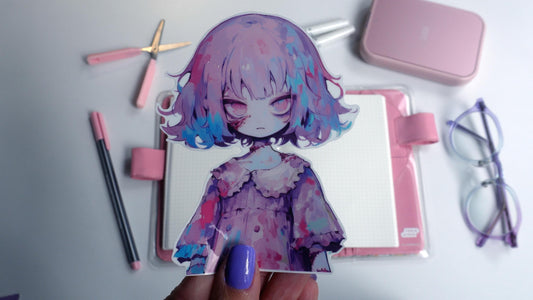 A hand with purple nails holds a sticker of an anime character with purple and blue hair, wearing a ruffled dress. Background items include a pink notebook, pens, scissors, a case, and glasses on a white surface.