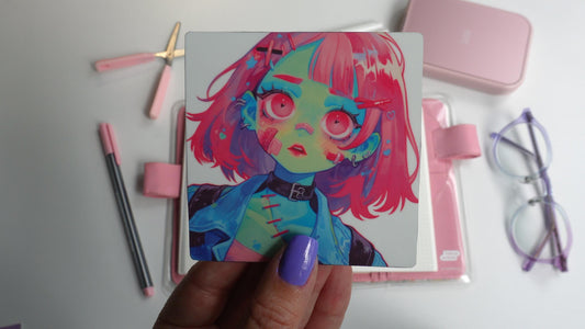 A hand with purple nails holds a square sticker of an anime character with pink hair, green skin, large pink eyes, and a choker. The background includes a pink notebook, pens, scissors, a pink case, and purple-rimmed glasses on a white surface.