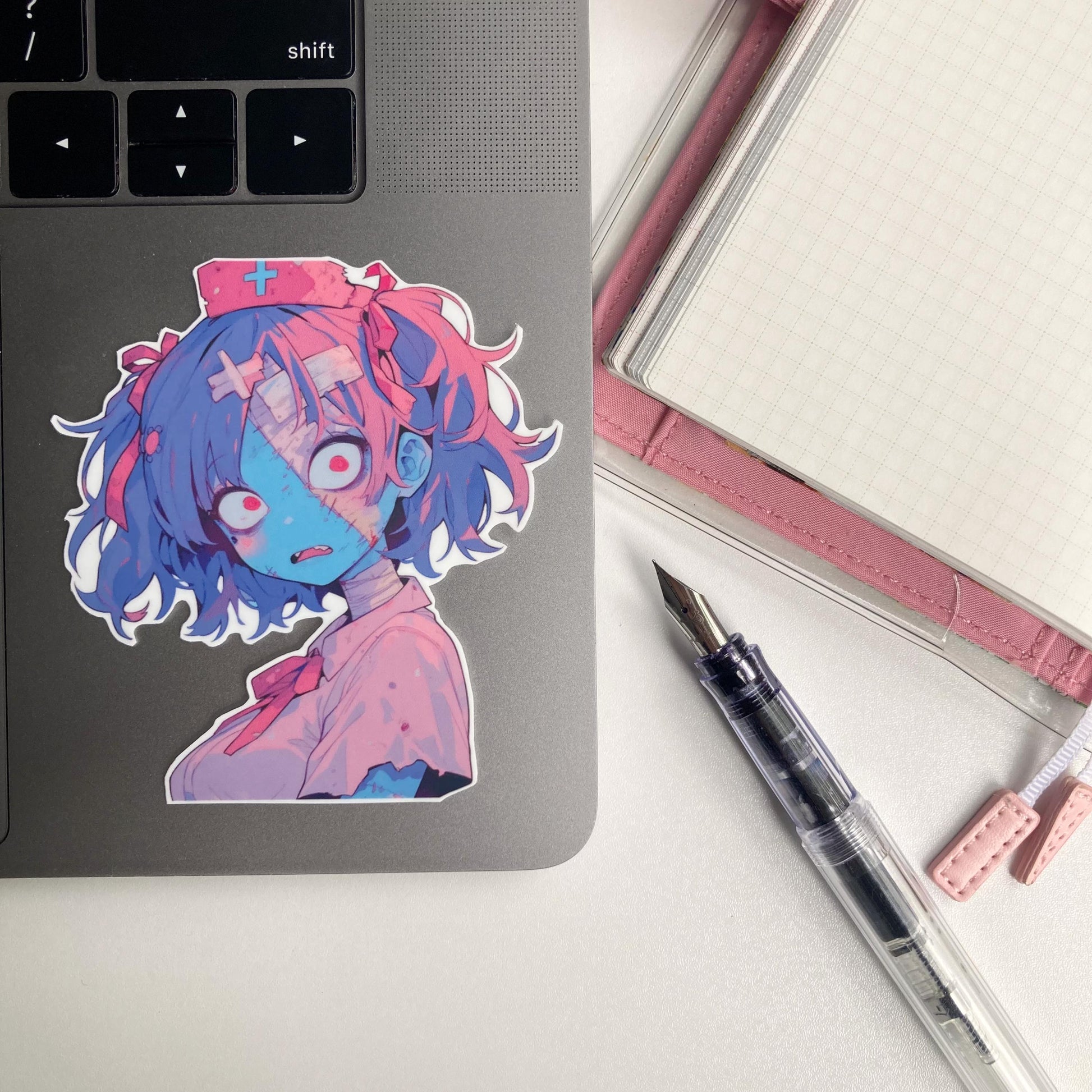 A sticker of a zombie nurse character with blue skin, pink hair, and a shocked expression is placed on a gray laptop. To the right are a clear fountain pen, a pink notebook, and its page marker. The laptop keyboard&#39;s &quot;shift&quot; key is partially visible.