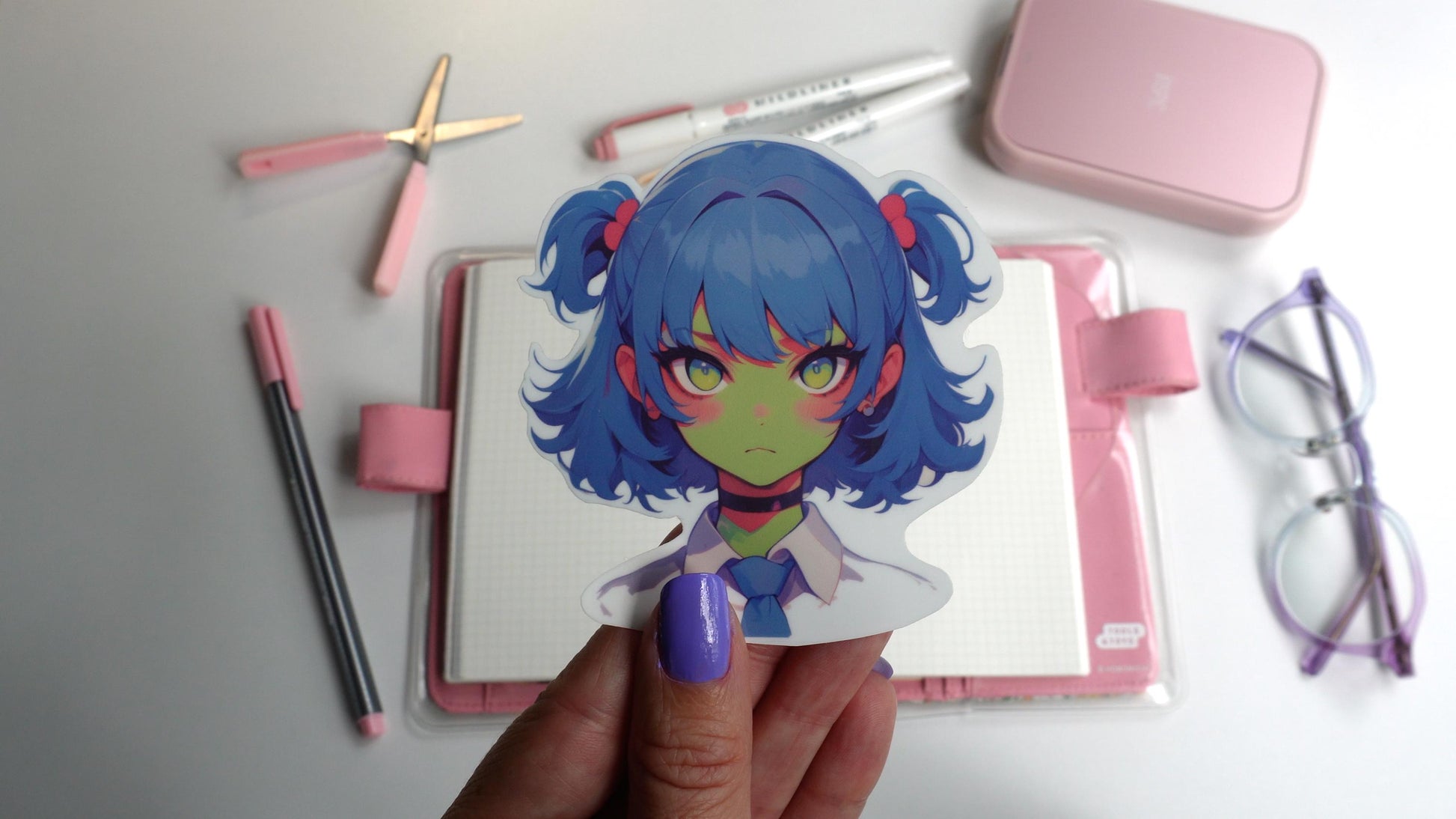 A sticker of an anime-style character with blue twin-tails, pink hair ties, green skin, and yellow eyes, wearing a white shirt, blue tie, and black choker. The sticker is held up by a hand with purple-painted nails.