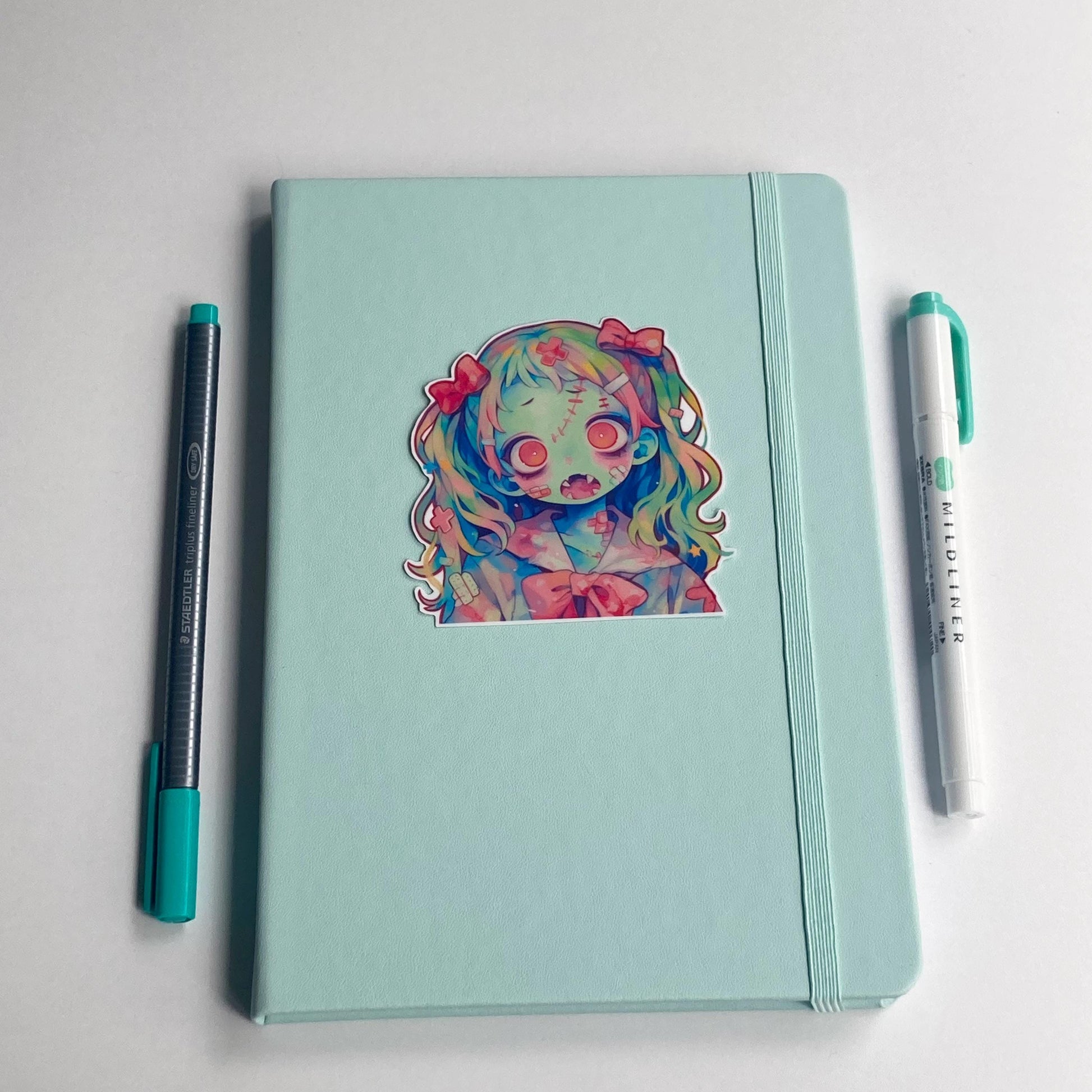 Sticker of an anime zombie kid with rainbow hair, pink bows, greenish-blue skin, and pink eyes on a mint notebook. The character features stitches, bandages, and a pink bow on her outfit. A teal pen and matching highlighter are placed on either side