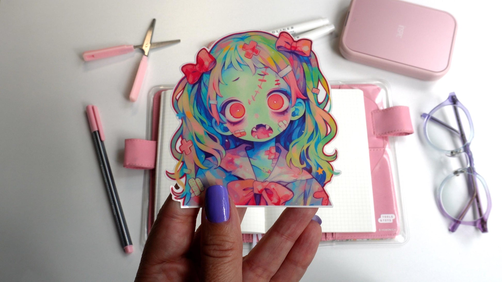 A sticker of an anime-style zombie girl with multicolored hair, pink bows, and a greenish-blue skin tone. She has large pink eyes, an open mouth with sharp teeth, and bandages. The sticker is held up by a hand with purple-painted nails.