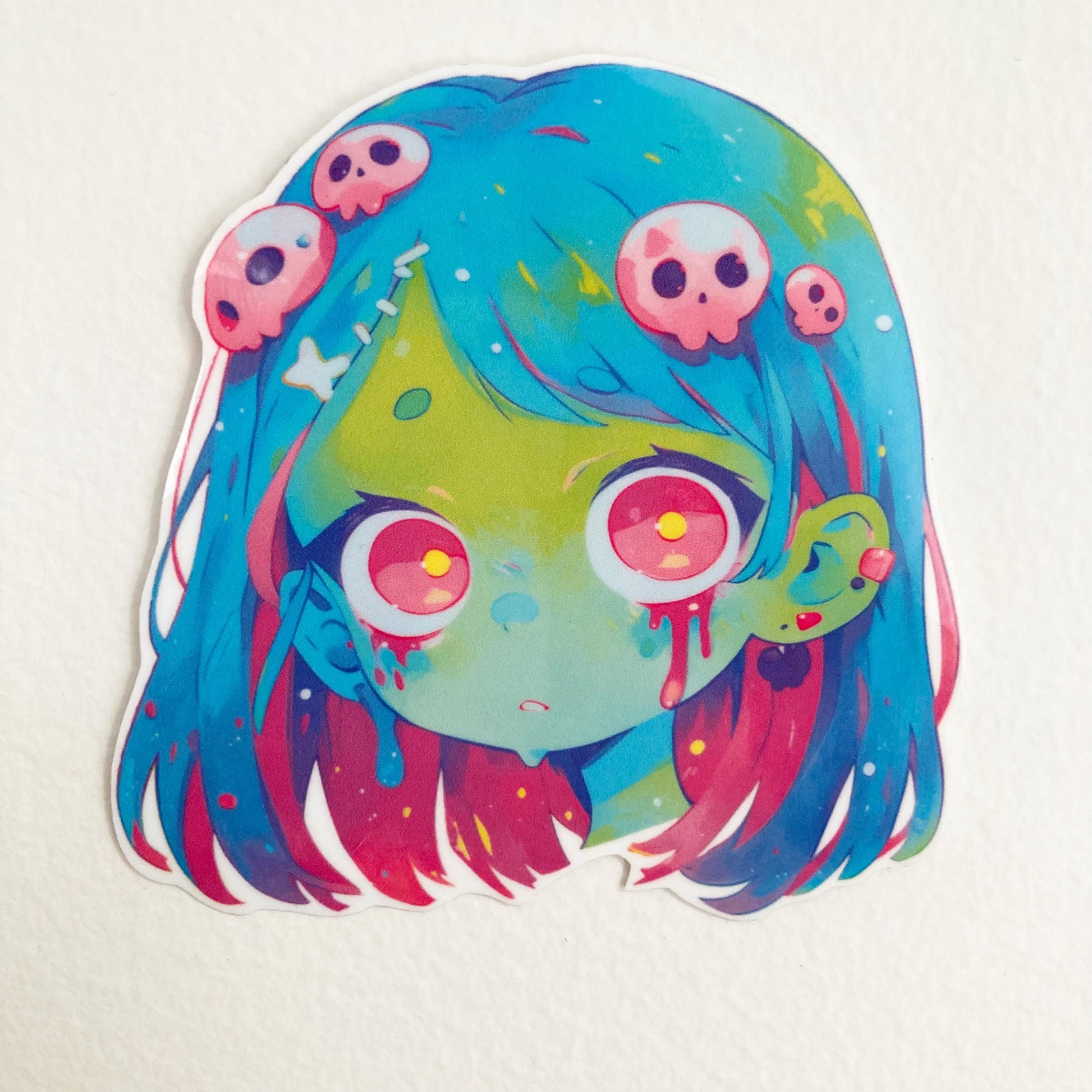 Sticker of an anime-style zombie girl with blue and pink hair, green skin, and large pink eyes. Blood drips from her eyes and ear, with small pink skulls in her hair. Set against a plain background.