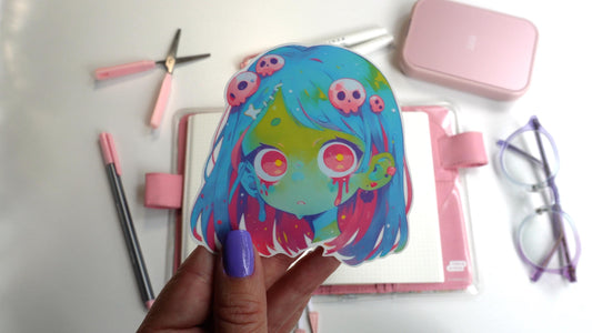 Sticker of an anime-style zombie girl with blue and pink hair, green skin, and large pink eyes. Blood drips from her eyes, and small pink skulls decorate her hair. The sticker is held up by a hand with purple-painted nails.