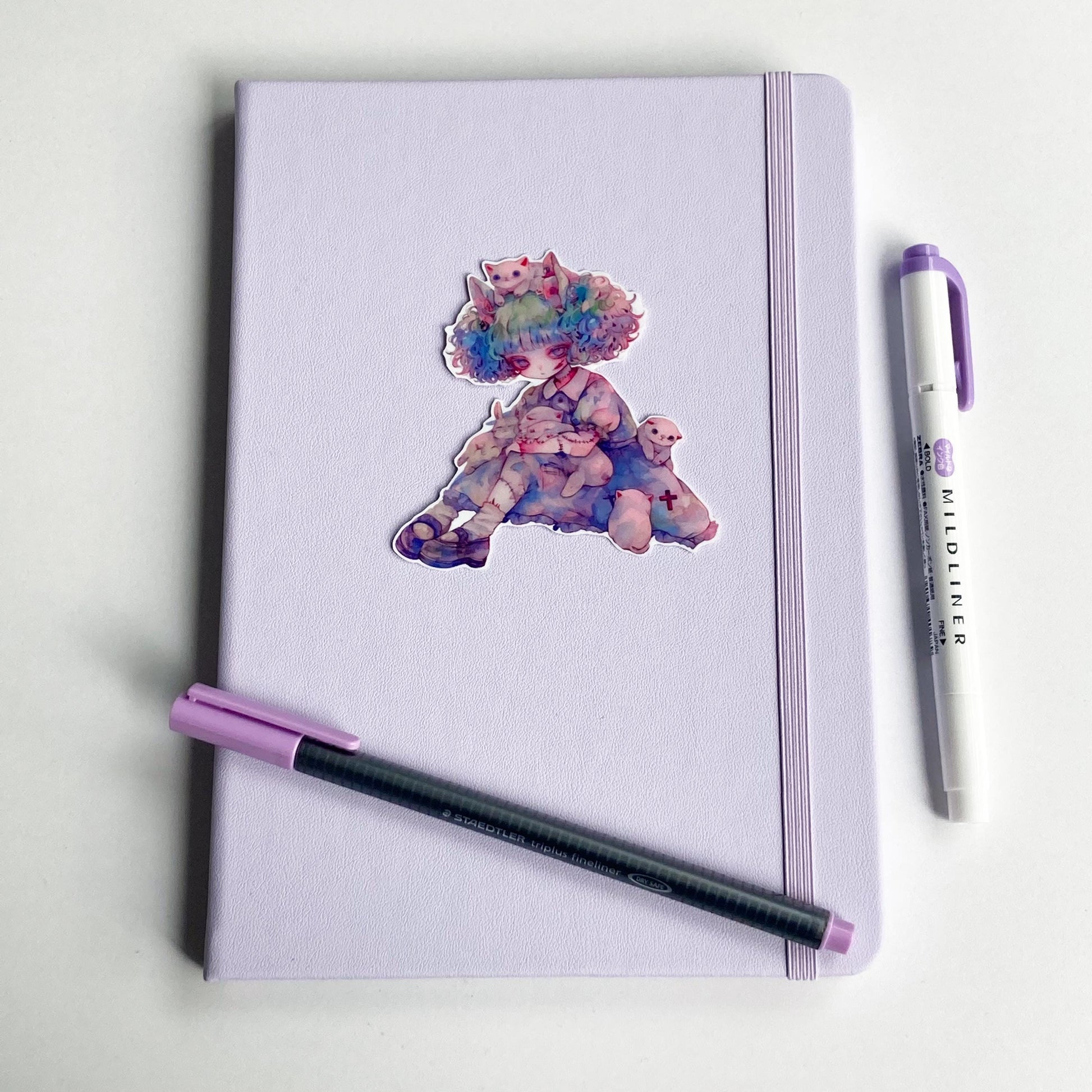 Sticker of an anime character with pastel curly hair sitting with white cats and bunnies, wearing a blue dress with stitched limbs. The sticker is placed on a lavender notebook, with a purple pen and matching highlighter nearby.