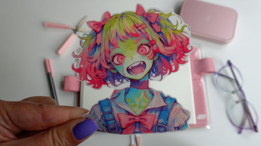 A hand with purple nails holds a colorful sticker of an anime-style zombie girl with green skin, pink and green hair, wide red eyes, and fangs. The background features a pink notebook, pen, scissors, and glasses on a white surface.