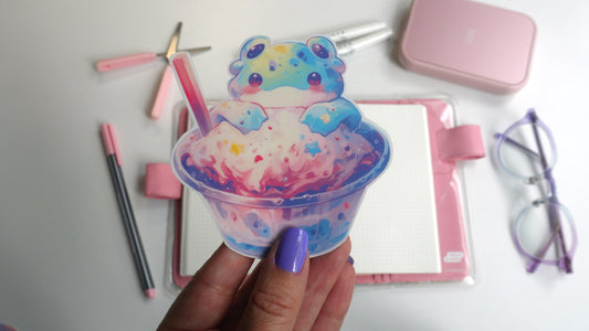 A hand with lavender nails holds a colorful sticker of a cute alien frog sitting in a cup of pastel pink and blue ice cream. The background includes pink stationery, a notebook, and a pair of clear glasses with purple accents.