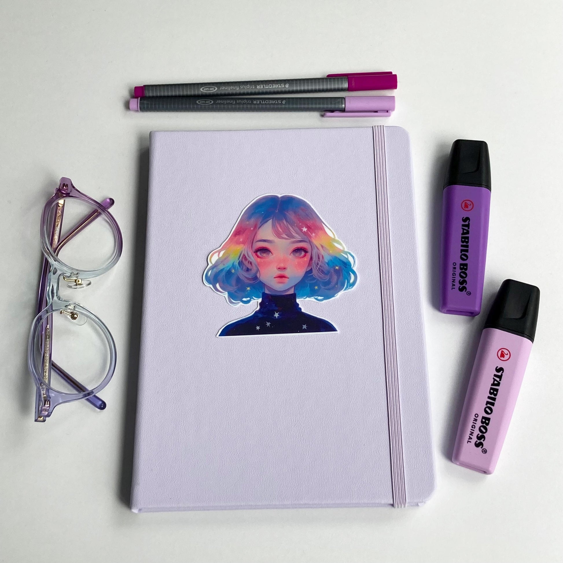 A lavender notebook with a sticker of a person with pastel rainbow hair is surrounded by purple-rimmed glasses, two highlighters, and two pens in shades of purple and pink. The items are neatly arranged on a white surface.