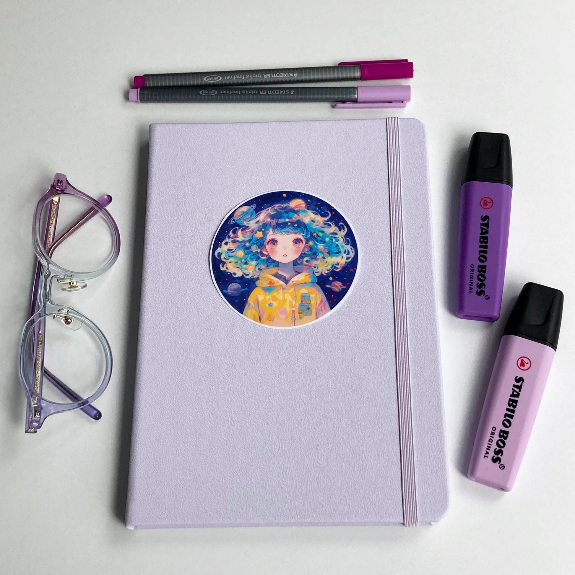 A lavender notebook with a circular sticker of a person with colorful, curly hair wearing a yellow space-themed hoodie is surrounded by purple-rimmed glasses, two highlighters, and two pens in shades of purple and pink.
