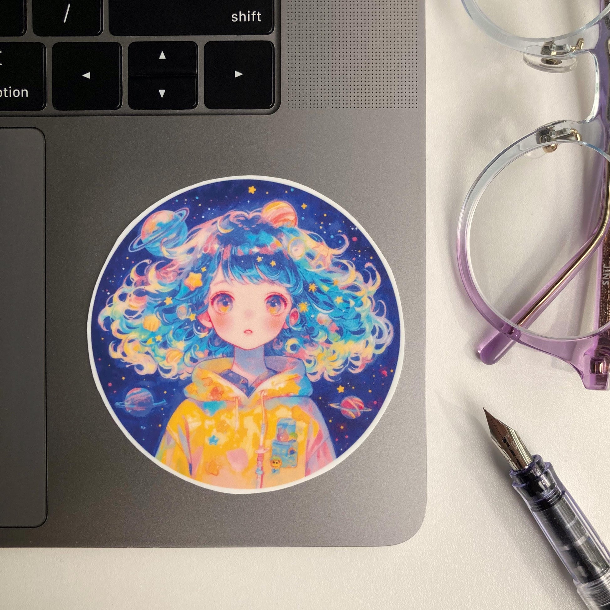 A circular sticker of a person with colorful, curly hair wearing a yellow space-themed hoodie is placed on a laptop. Nearby are purple-rimmed glasses and a clear fountain pen.
