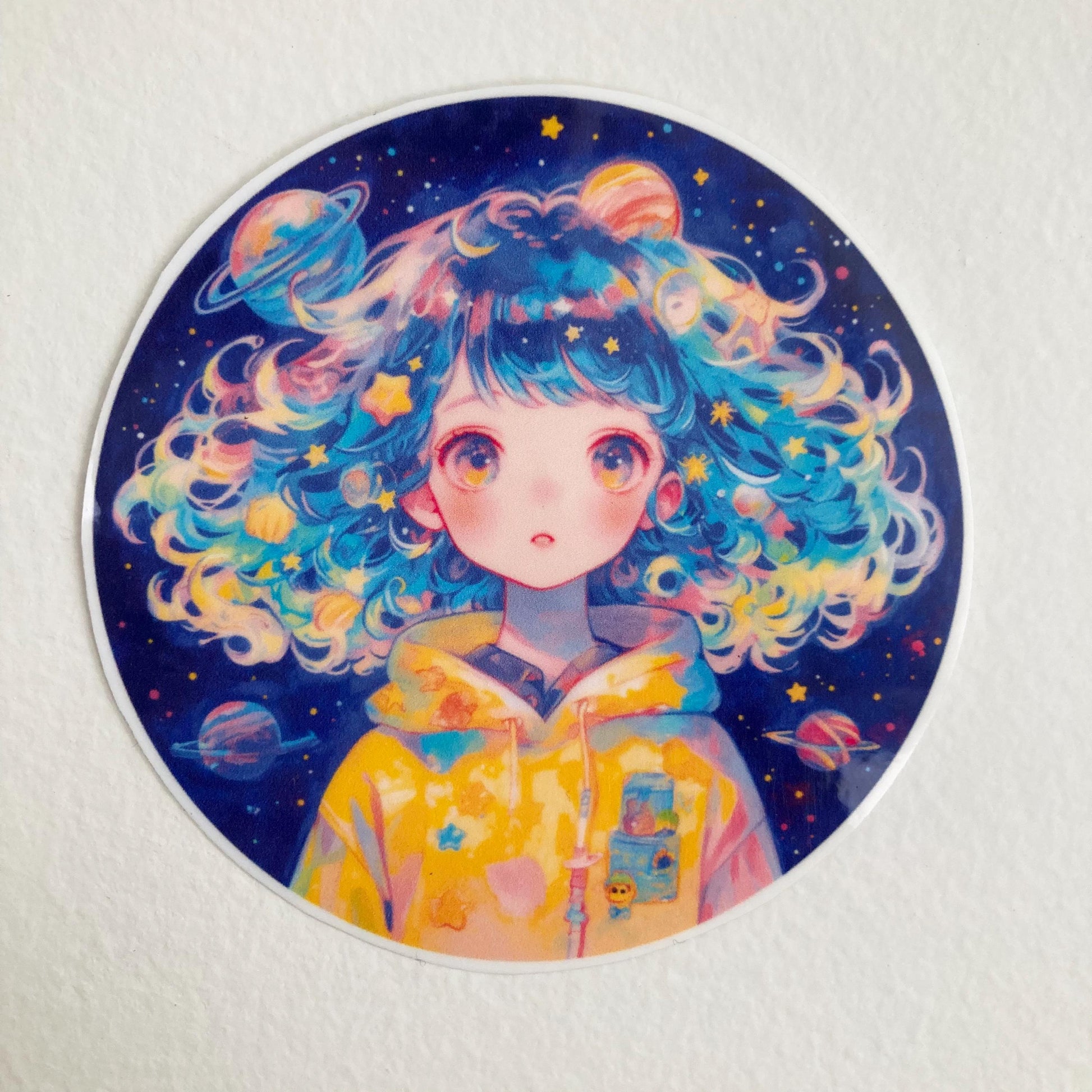 A circular sticker featuring a person with curly, multicolored hair surrounded by stars and planets. They wear a yellow hoodie with space-themed designs, set against a dark, starry background.