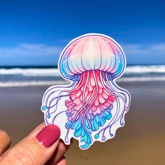A hand with pink nails holds a colorful jellyfish sticker with shades of pink, blue, and purple. The background shows a sandy beach with the ocean and a clear blue sky, highlighting the intricate details of the jellyfish against the coastal scene.