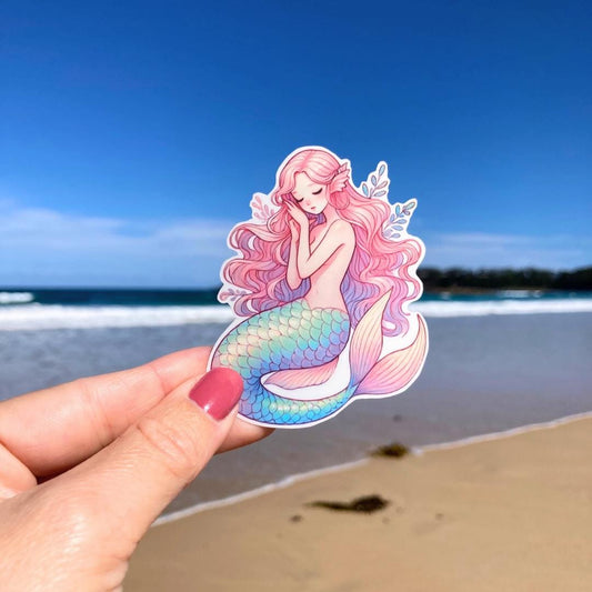 A hand holds a sticker of a mermaid with long pink hair and a blue-green tail, sitting gracefully. The background shows a sandy beach with the ocean and a clear blue sky, highlighting the mermaid&#39;s delicate details against the coastal scene