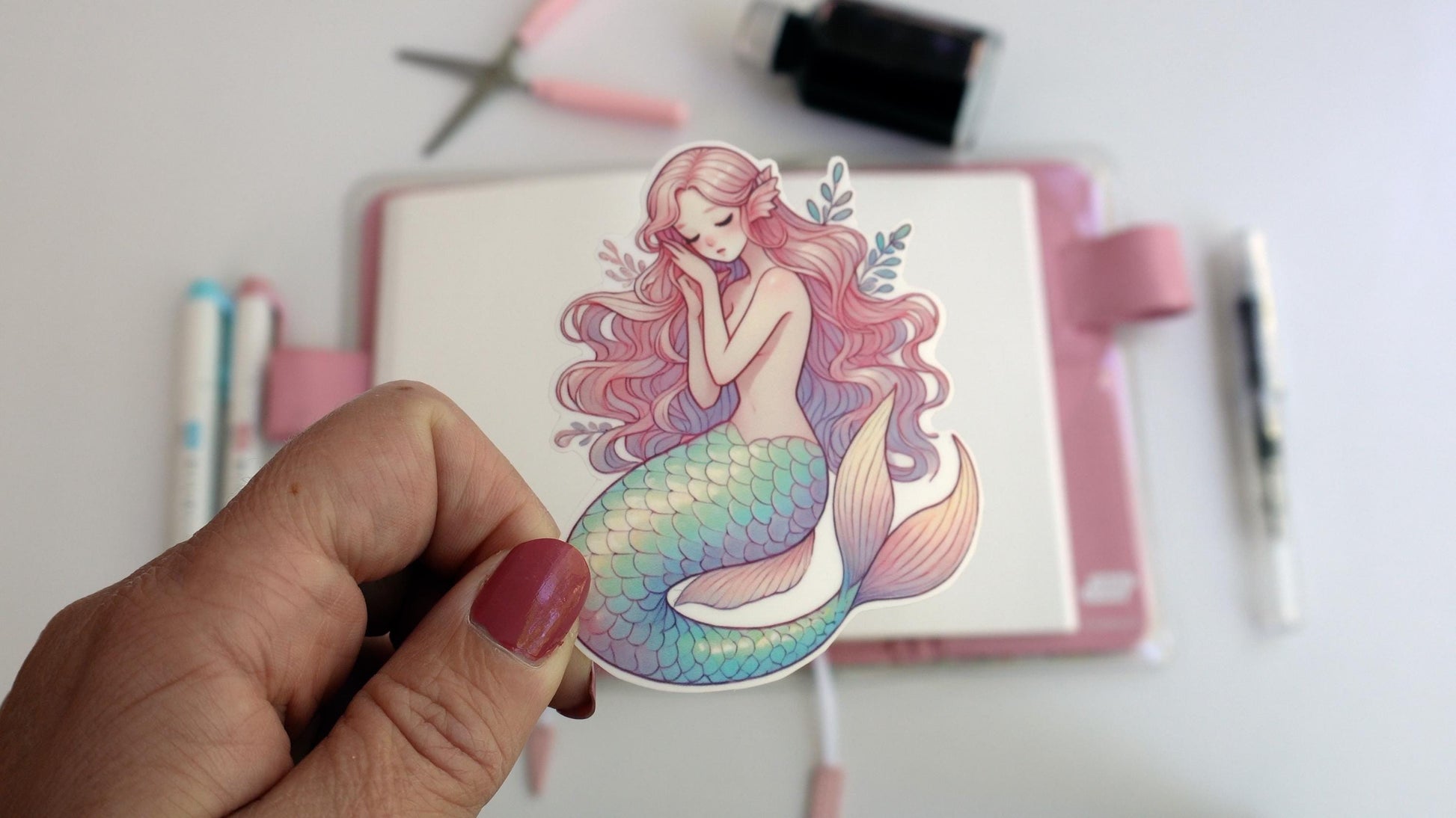 A hand holds a sticker of a serene pastel mermaid with long pink hair and a teal, lavender, and yellow scaled tail. The background shows a desk setup with pens, scissors, and an ink bottle, creating a creative workspace atmosphere.