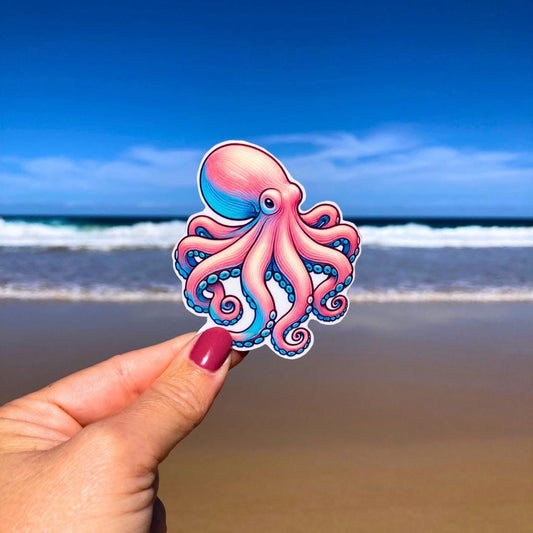 A hand with pink nails holds a colorful octopus sticker with pink and blue hues. The background shows a sandy beach with waves and a clear blue sky, highlighting the octopus&#39;s swirling tentacles against the serene coastal scene.
