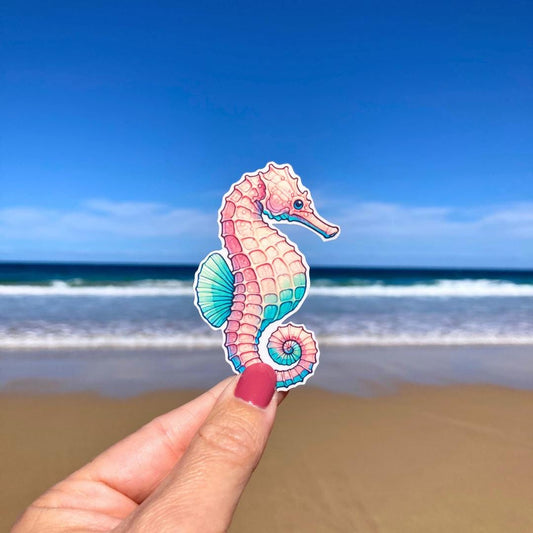 A hand with pink nails holds a colorful seahorse sticker with pink, blue, and green tones. The background shows a sandy beach with waves and a bright blue sky, highlighting the seahorse&#39;s intricate details against the serene beach scene.