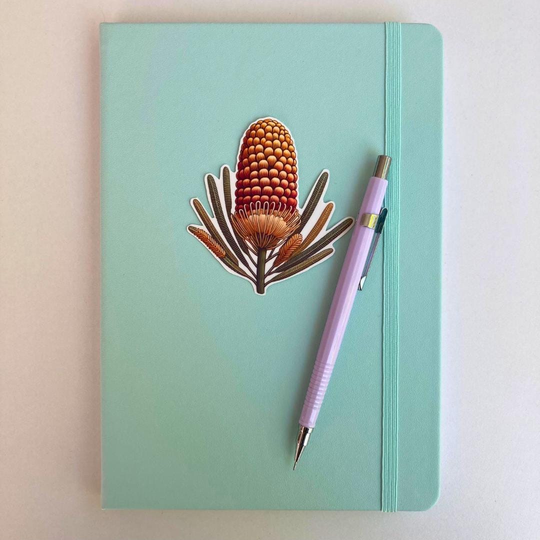 A light turquoise notebook with an elastic strap features a sticker of an orange and yellow banksia flower with slender leaves. A lavender mechanical pencil is placed diagonally on the notebook, which rests on a white surface.