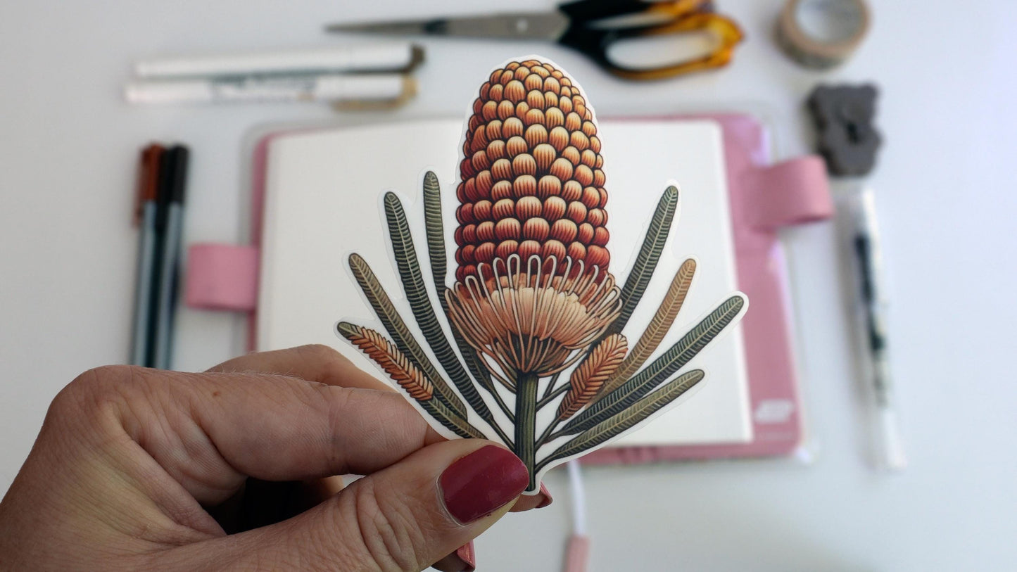 A hand with pink nail polish holds a sticker of an orange and yellow banksia flower with slender leaves. In the background, an open notebook with a pink cover sits on a white surface, along with scattered pens, scissors, and a roll of tape.