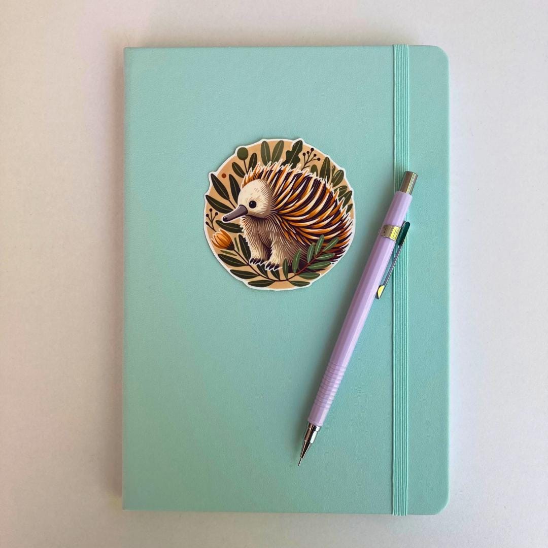 A mint green notebook with a round sticker of an echidna surrounded by leaves and an orange flower. The echidna has brown, white, and yellow spines. A lavender mechanical pencil is placed diagonally on the notebook, near the sticker.