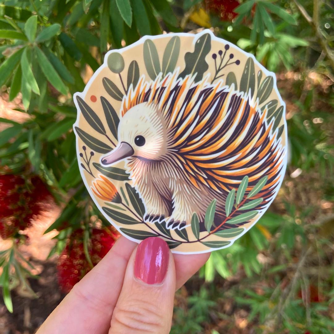 A hand with pink nail polish holds a round sticker of an echidna with brown, white, and yellow spines surrounded by green leaves and small orange flowers. In the background, blurred green leaves and red bottlebrush flowers are visible.