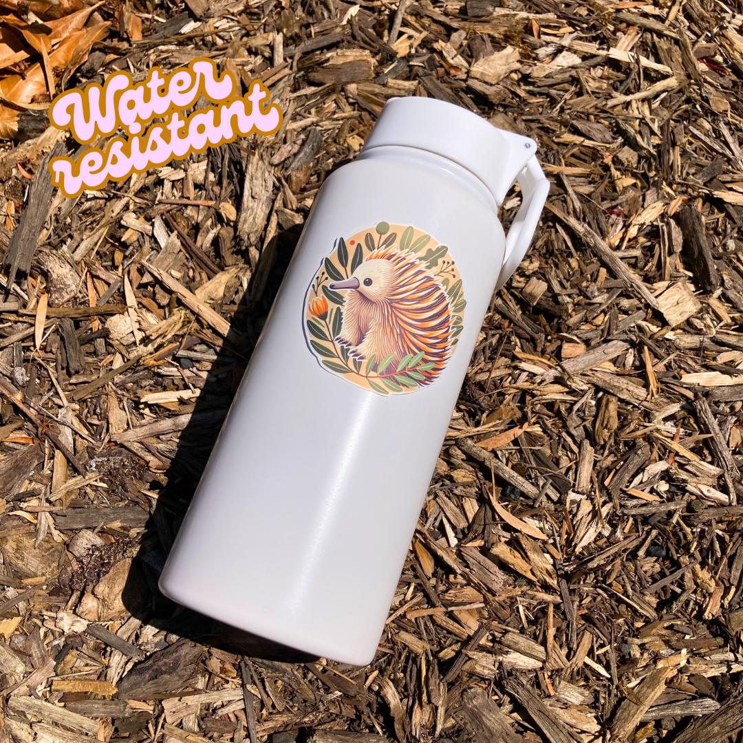 A white water bottle with a round sticker of an echidna surrounded by green leaves and small orange flowers rests on wood chips in bright sunlight. An orange &quot;Water resistant&quot; label is in the top left corner