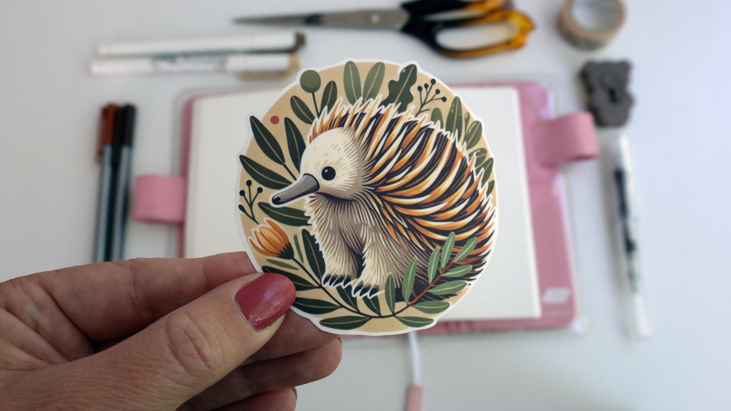 A hand holds a circular sticker featuring an echidna with orange, yellow, and white spines. The echidna is surrounded by green leaves and an orange flower. In the background are crafting supplies like scissors, pens, and a notebook.
