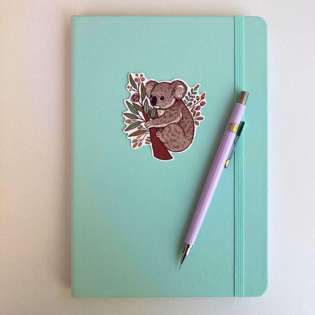 A light turquoise notebook with an elastic strap features a sticker of a koala hugging a tree trunk with eucalyptus leaves and branches. A lavender mechanical pencil is placed diagonally on the notebook, which rests on a white surface.