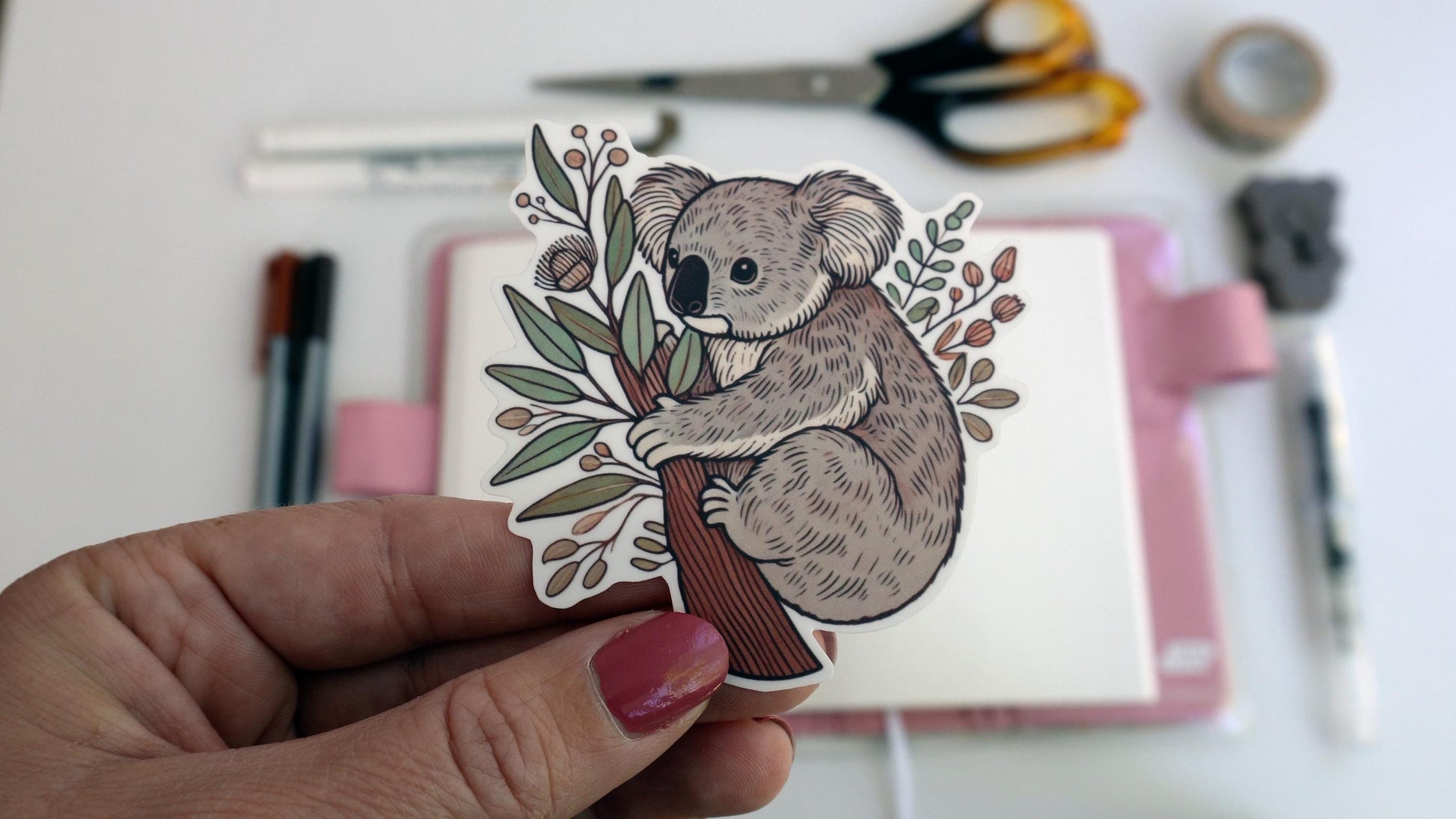A hand with pink nail polish holds a sticker of a koala hugging a tree trunk surrounded by eucalyptus leaves and branches. In the background, an open notebook with a pink cover sits on a white surface along with scattered pens, scissors, and tape.