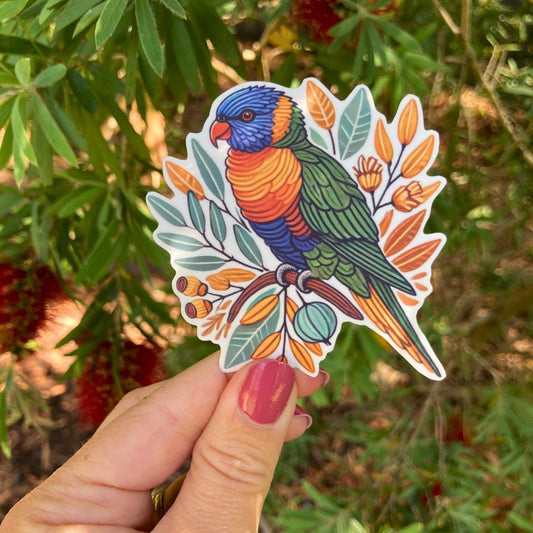 A hand holds a sticker of a colorful rainbow lorikeet perched on a branch with leaves and buds. The lorikeet has vibrant blue, green, orange, and yellow feathers. In the background, green leaves and red bottlebrush flowers are visible
