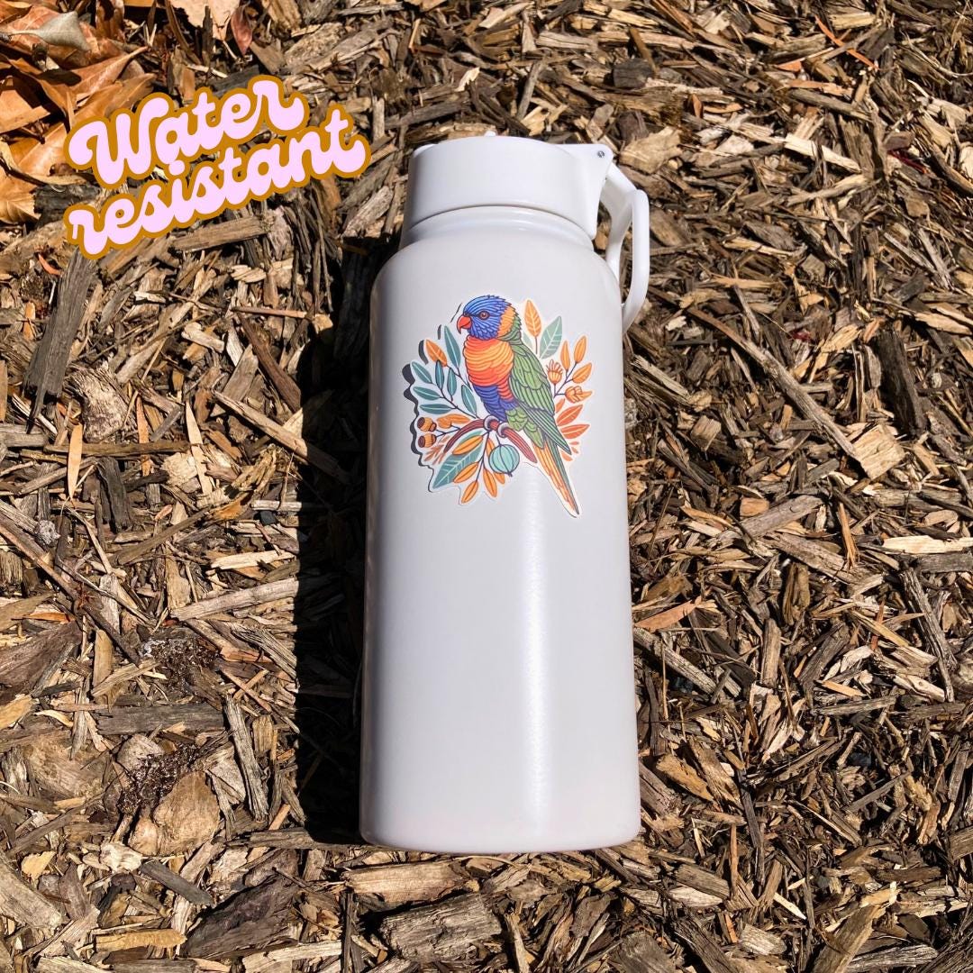 A white water bottle with a sticker of a colorful rainbow lorikeet perched on a branch with leaves and buds lies on wood chips in bright sunlight. An orange &quot;Water resistant&quot; label is in the top left corner