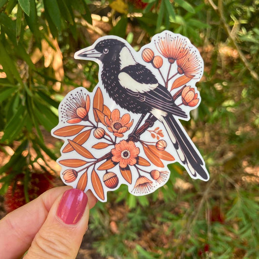 A hand with pink nail polish holds a sticker of a black-and-white magpie perched on a branch with orange flowers and leaves. In the background, green leaves and red bottlebrush flowers are visible, blending with the natural theme of the sticker.