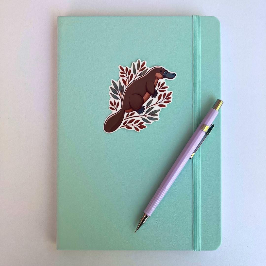 A sticker of a cartoon platypus with leaves is placed on a closed light turquoise notebook. A lavender mechanical pencil rests diagonally on the notebook, which has an elastic strap on the right side. The background is a plain white surface.