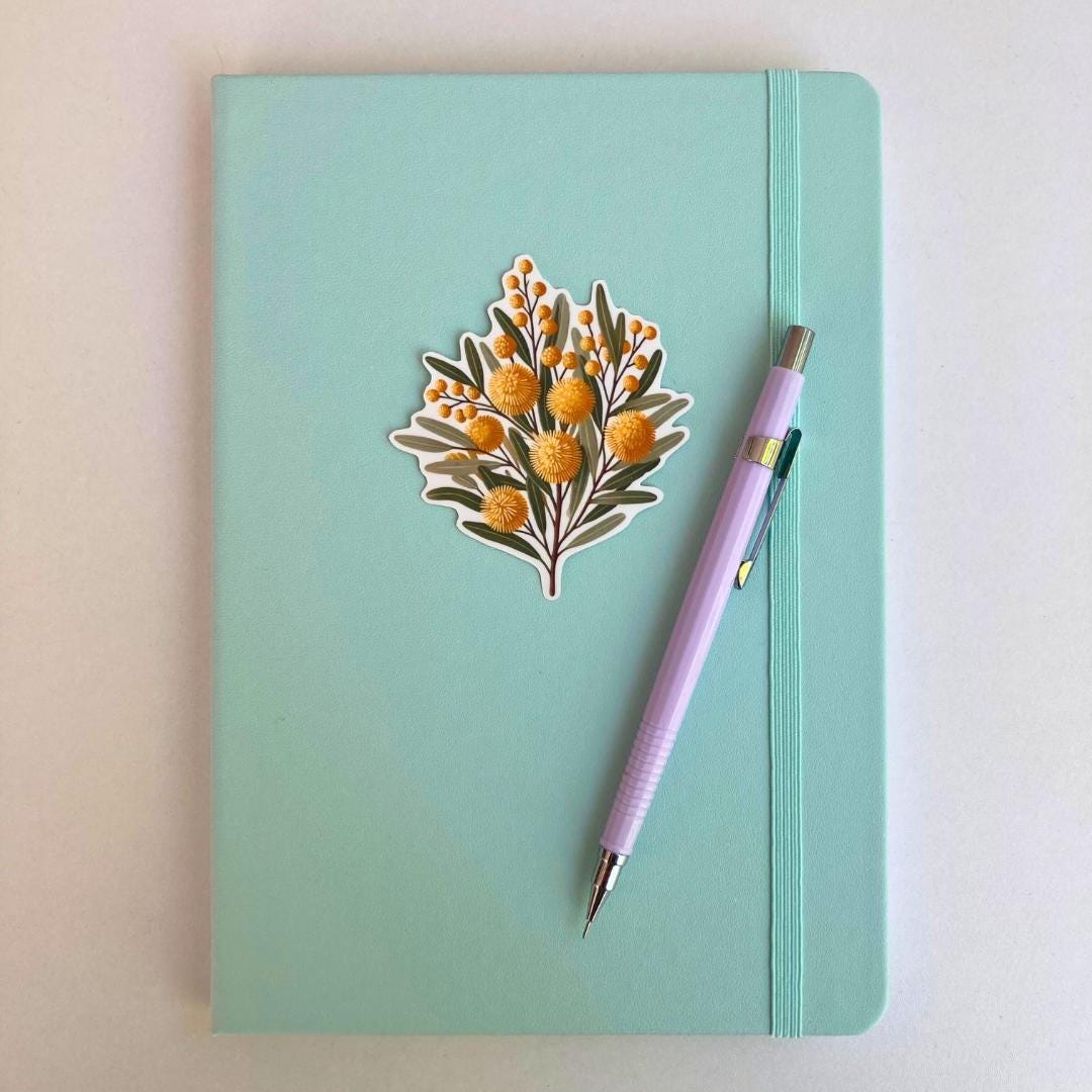 A teal notebook with a sticker of yellow wattle flowers in the center. The design features round, fluffy yellow blossoms and green leaves. A lavender mechanical pencil with a silver clip rests diagonally on the right side of the notebook.