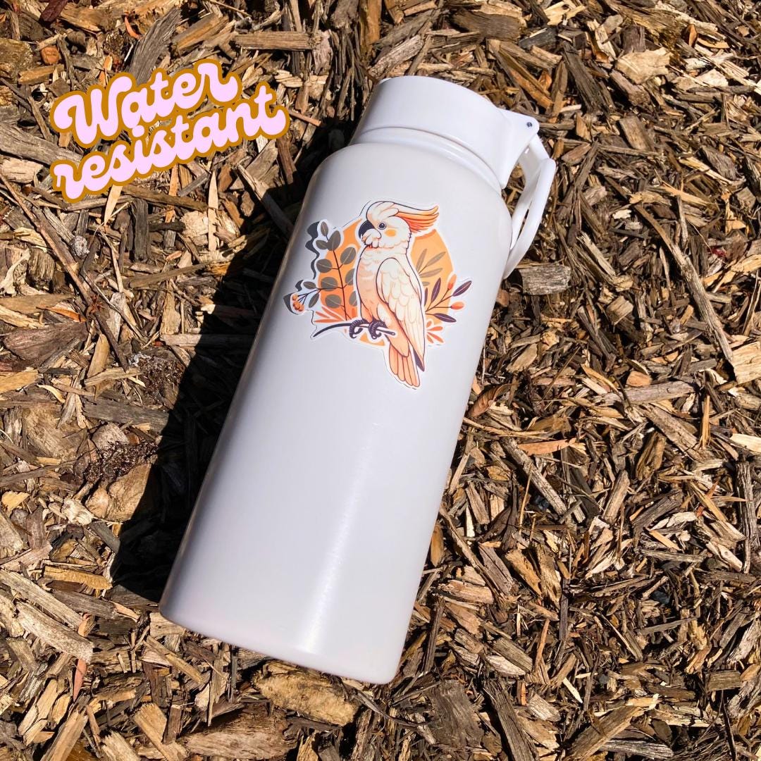 A white water bottle lies on wood chips, featuring a sticker of a white cockatoo with an orange crest perched on a branch, surrounded by leaves. The phrase &quot;Water resistant&quot; is displayed in stylized orange text in the upper left corner of the image.