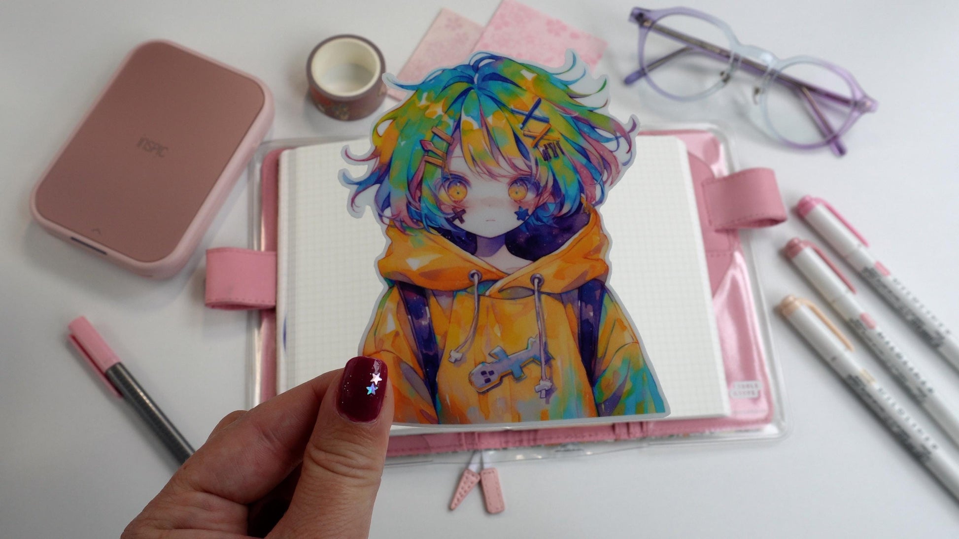 A hand holds a anime sticker with a character in a yellow hoodie and multicolored hair over a pink planner. Surrounding items include purple glasses, pink markers, washi tape, and a pink printer. The hand has pink nail polish with tiny star decals.