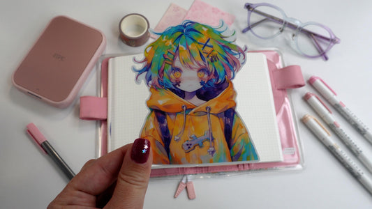 A hand holds a anime sticker with a character in a yellow hoodie and multicolored hair over a pink planner. Surrounding items include purple glasses, pink markers, washi tape, and a pink printer. The hand has pink nail polish with tiny star decals.