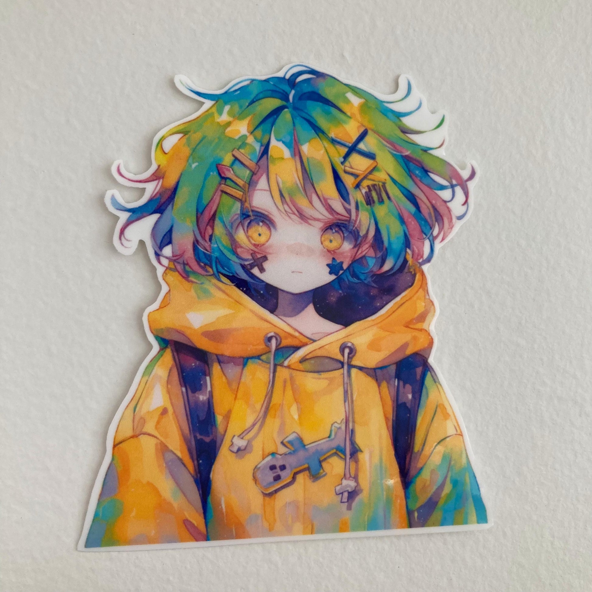 A colorful anime sticker featuring a character with vibrant multicolored hair and bright yellow eyes, wearing a yellow hoodie adorned with a crossbone design. The sticker is placed on plain, light background, highlighting its vivid colors and details
