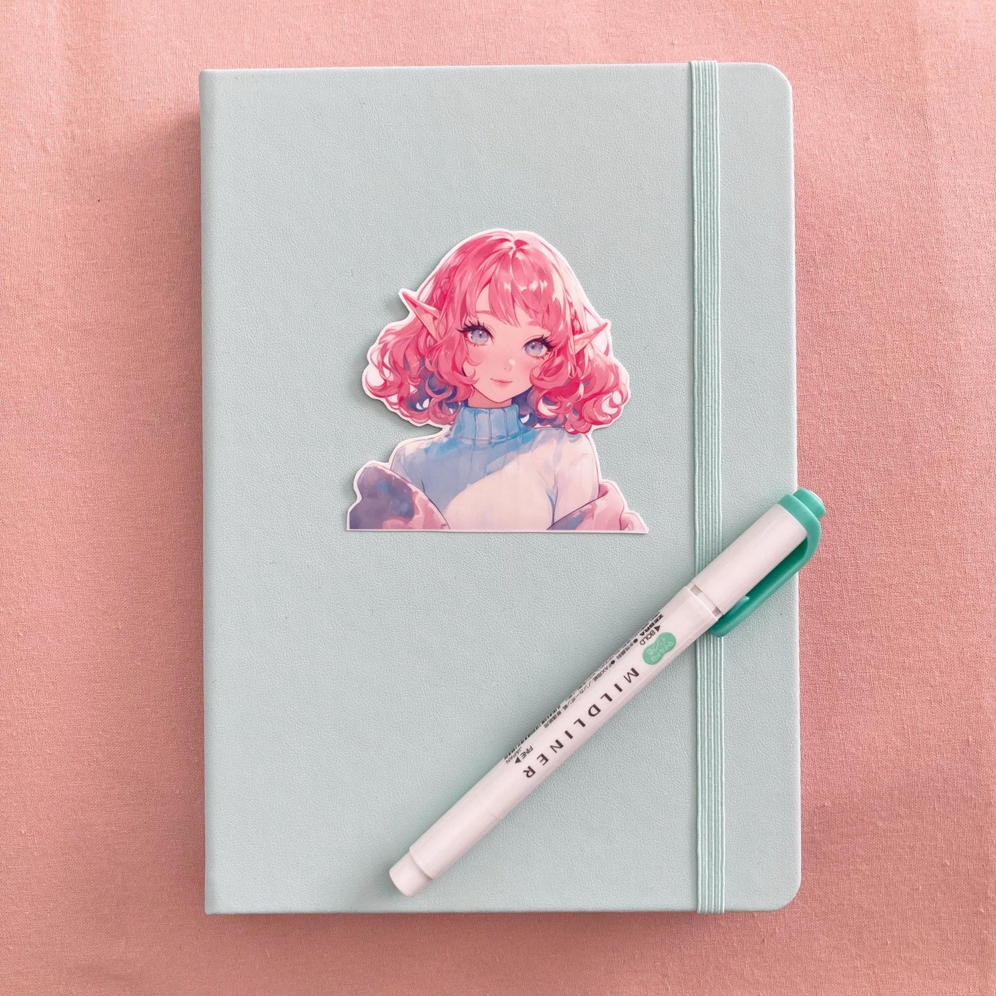 A pastel green notebook with an anime sticker featuring a character with curly pink hair, elf ears, and bright eyes wearing a turtleneck. A green Mildliner highlighter rests diagonally on the notebook, set against a soft pink fabric background.
