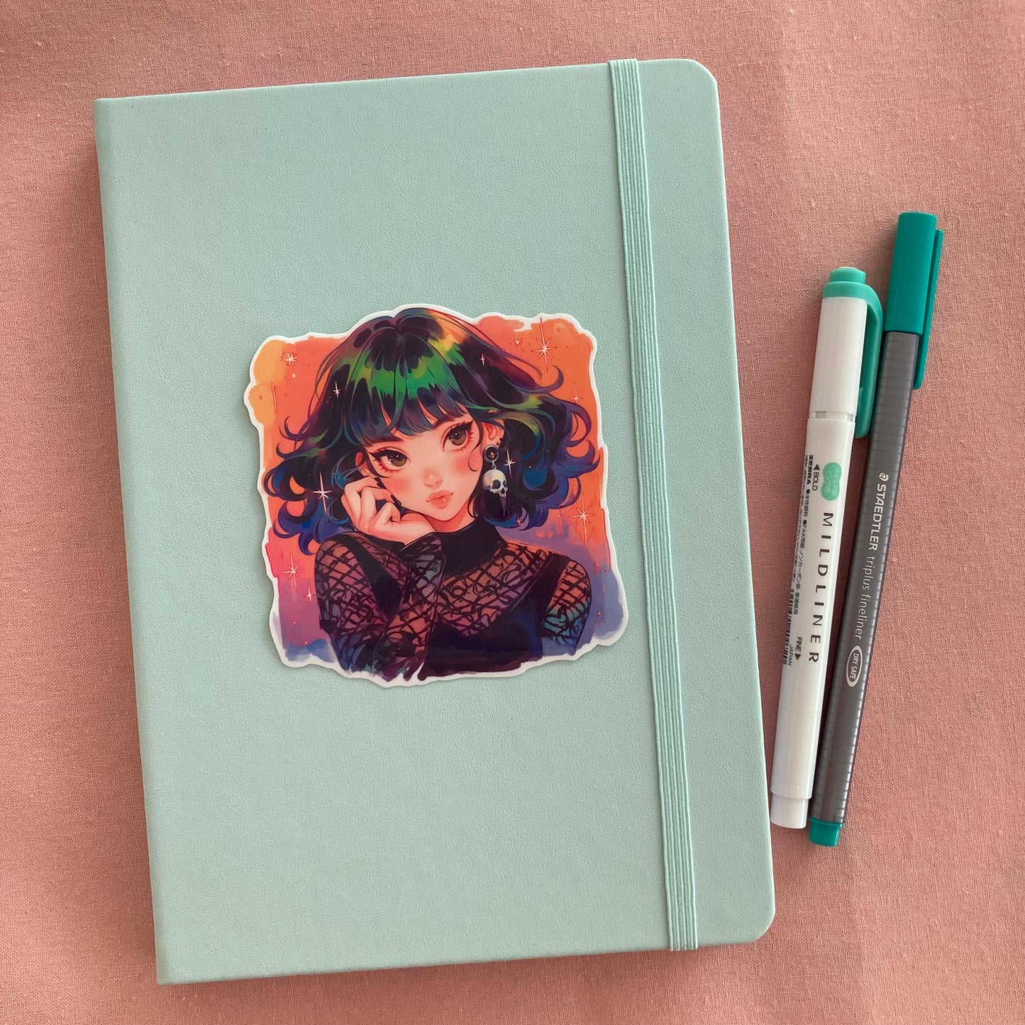 A pastel green notebook with a colorful anime sticker of a girl with rainbow hair, star accents, and a skull earring in a gothic mesh outfit. Next to the book are a teal Mildliner highlighter and a green Staedtler pen, set against a soft pink fabric