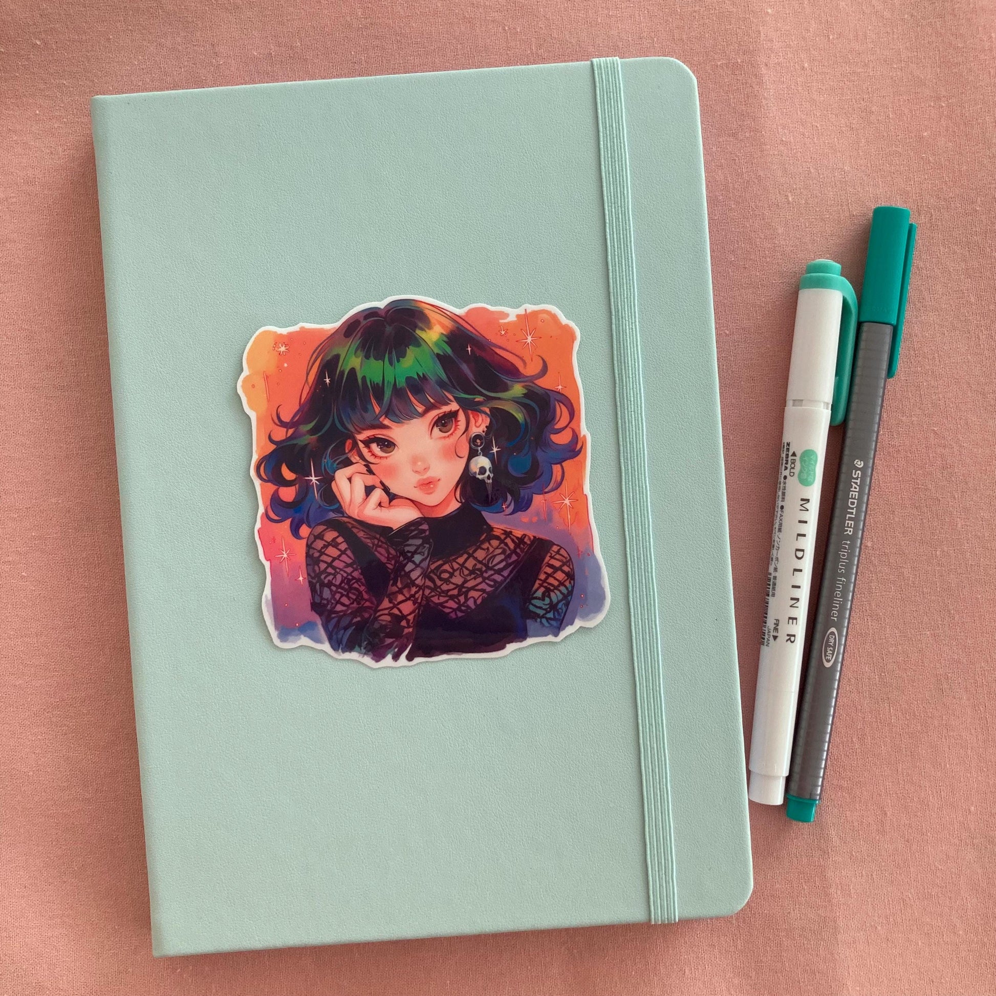 A pastel green notebook with a colorful anime sticker of a girl with rainbow hair, star accents, and a skull earring in a gothic mesh outfit. Next to the book are a teal Mildliner highlighter and a green Staedtler pen, set against a soft pink fabric