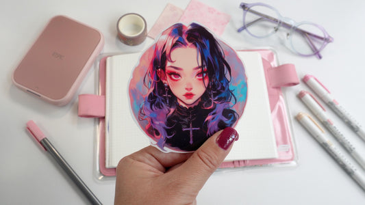 A hand holding a colorful anime sticker featuring a girl with dark hair streaked in pink and blue, wearing a cross necklace. The background includes a pink planner, purple glasses, markers, washi tape, and a pink printer, adding to the aesthetic.