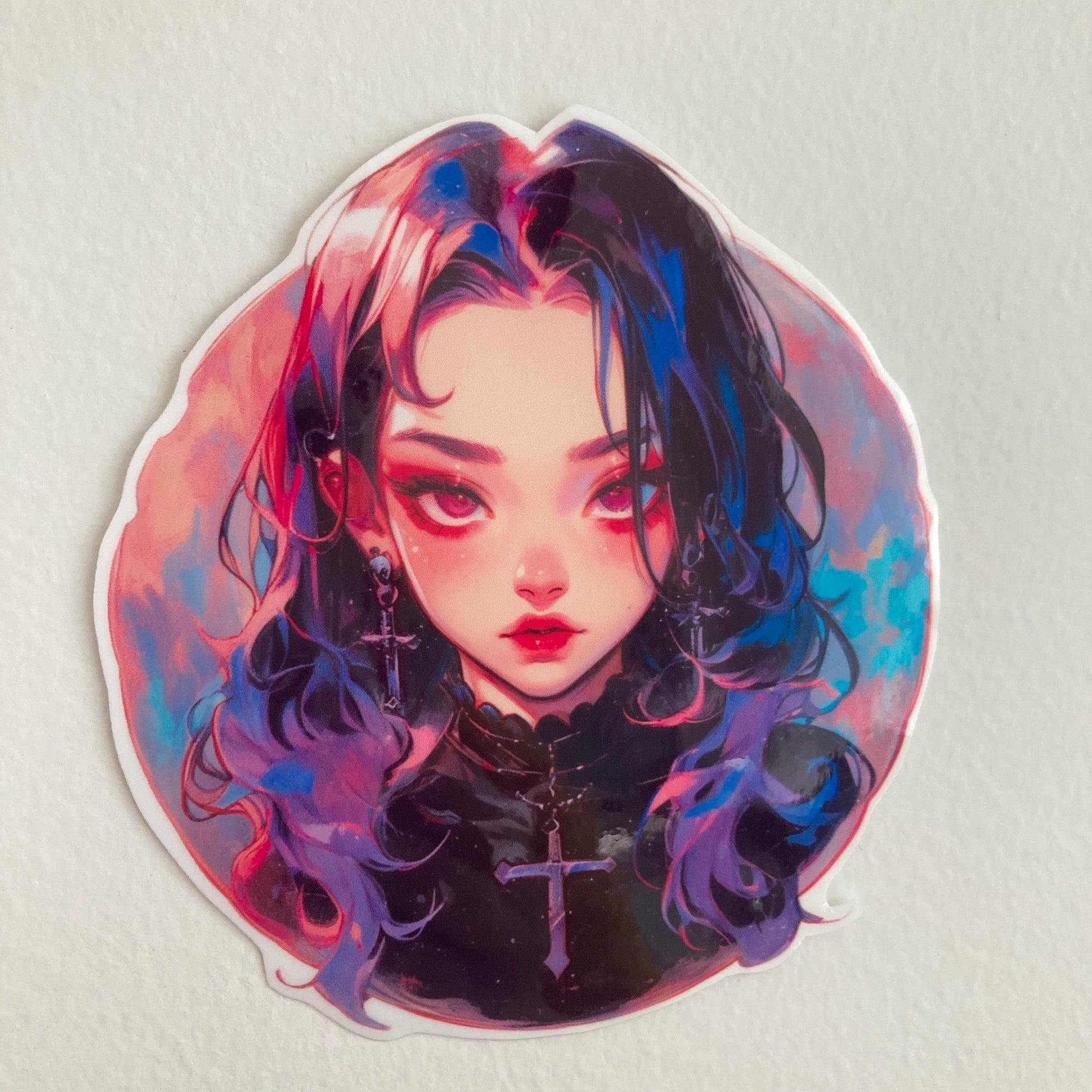A close-up of an anime-style sticker featuring a girl with long, dark hair with pink and blue highlights, wearing a black top and cross earrings and necklace. The background is a blend of pink and blue hues. Sticker is placed on a light surface.