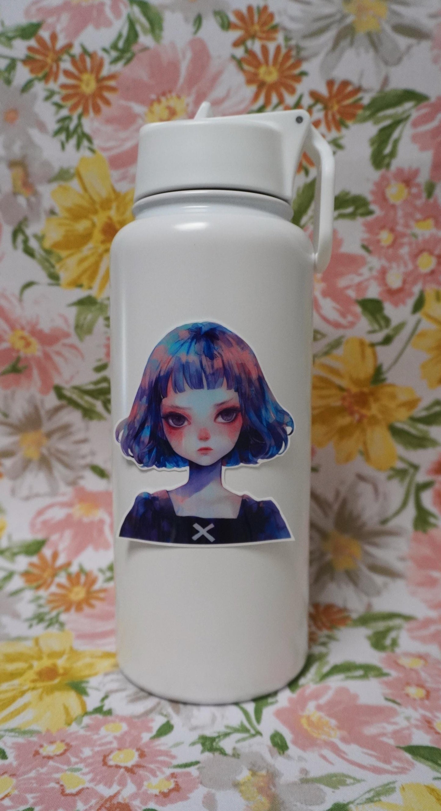 A white water bottle decorated with a sticker of a girl with blue and purple hair. The background is a floral fabric with large yellow, pink, and orange flowers, along with green leaves.